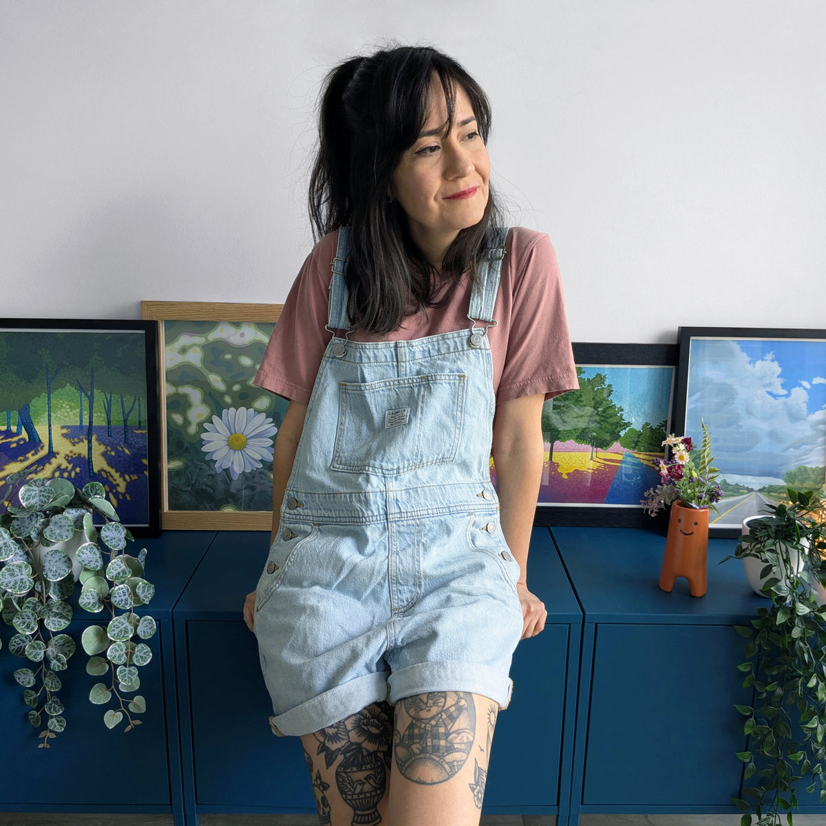 An artist sitting in front of a wall with framed paintings and plants